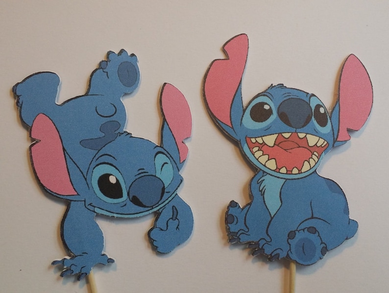 Puede incluir: Dos adornos para tartas de Stitch azul con orejas rosas. Un Stitch est&aacute; de pie sobre su cabeza con el brazo levantado, el otro Stitch est&aacute; sentado con la boca abierta.