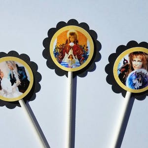 May include: Three black and gold round cupcake toppers with images from the movie Labyrinth. One topper features a close-up of the Goblin King, another features the Goblin King with a young girl, and the third features a young girl and a man in a suit.