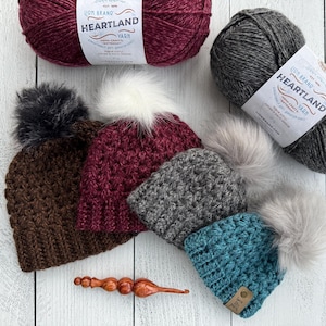 May include: Four crocheted hats with faux fur pom poms in brown, burgundy, gray, and teal. The hats are arranged in a row on a white wooden surface. Three skeins of Lion Brand Heartland yarn in burgundy, gray, and teal are also shown.