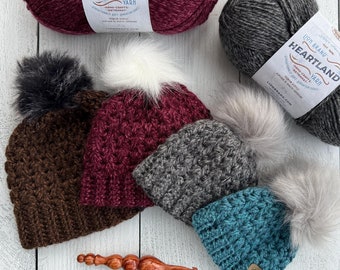 May include: Four crocheted hats with faux fur pom poms in brown, burgundy, gray, and teal. The hats are arranged in a row on a white wooden surface. Three skeins of Lion Brand Heartland yarn in burgundy, gray, and teal are also shown.