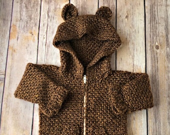 May include: A brown crocheted hooded jacket with bear ears. The jacket has a zipper closure and a ruffled hem.