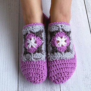 Puede incluir: Un par de zapatillas de crochet de color morado con un diseño floral gris y blanco.