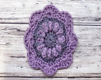 May include: A purple and grey crocheted flower coaster with a textured design.