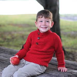 Puede incluir: Un niño pequeño con un suéter de punto rojo con botones marrones está sentado en un banco de madera sosteniendo una manzana roja.