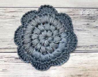 May include: A crocheted flower coaster in a blue and gray color gradient. The coaster is made with a circular design and has a textured surface.