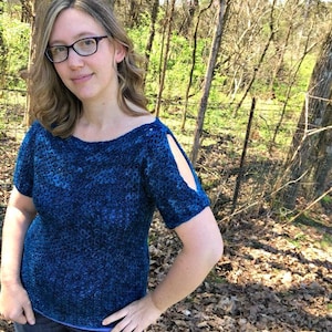 May include: A woman wearing a blue, textured, short-sleeved shirt with cut-out details on the shoulders. The shirt has a scoop neckline.