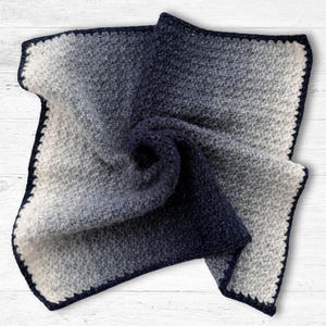 Könnte beinhalten: Eine gefaltete, quadratische Decke mit einem Farbverlauf von Hellgrau zu Dunkelmarineblau. Die Decke hat eine strukturierte Optik und einen dunkelblauen Rand.