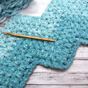 Könnte beinhalten: Nahaufnahme einer blauen Häkeldecke mit einer Holz-Häkelnadel darauf. Die Decke hat einen Farbverlauf in Blau und eine strukturierte, flauschige Oberfläche.