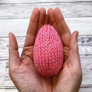 K&ouml;nnte beinhalten: Ein rosa gestricktes Ei, das in hohlen H&auml;nden gehalten wird. Das Ei ist mit einem strukturierten Strickmuster gefertigt.