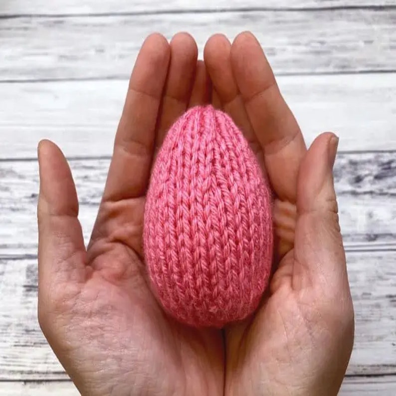 K&ouml;nnte beinhalten: Ein rosa gestricktes Ei, das in hohlen H&auml;nden gehalten wird. Das Ei ist mit einem strukturierten Strickmuster gefertigt.