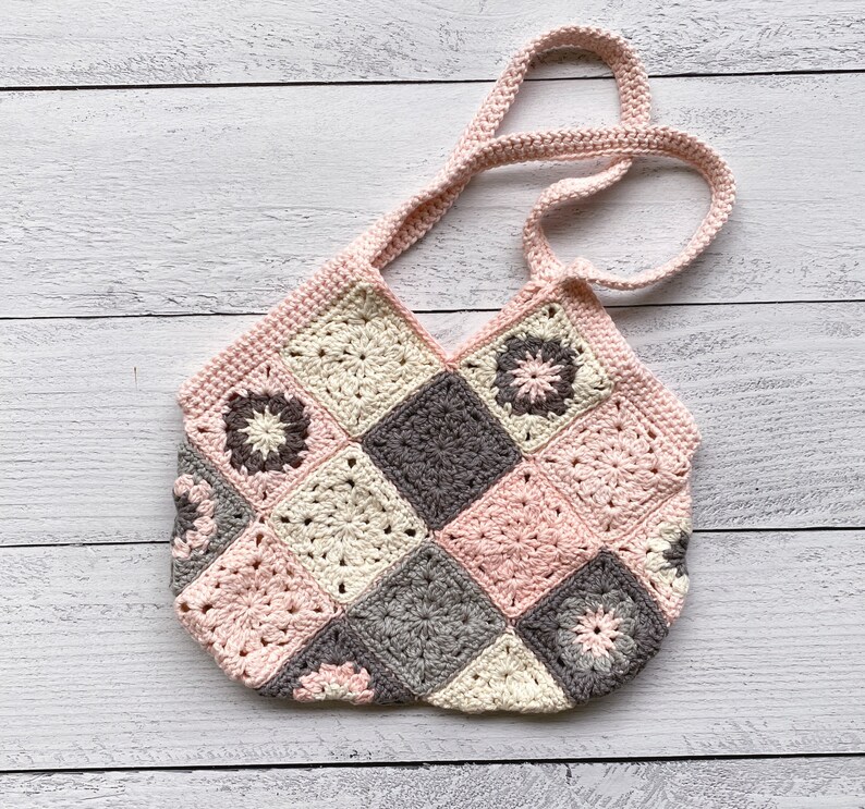 May include: A crocheted tote bag with a patchwork design featuring pink, cream, and gray squares. The bag has a single strap and is made with a granny square stitch.