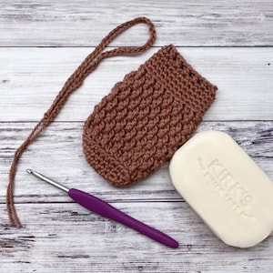 May include: A brown crocheted soap pouch with a long brown yarn tail. A purple crochet hook and a white bar of soap with the text "Kirk's Castile" are also pictured.