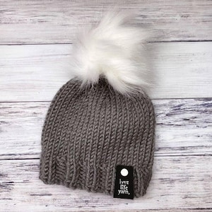 May include: A gray knit beanie with a large white faux fur pom-pom. The hat has a ribbed texture and a small black tag that reads "love life yarn." The hat is displayed on a light wood background.
