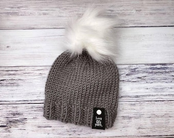 May include: A grey knitted beanie hat with a large white faux fur pom-pom. The hat has a ribbed texture and a small black tag that reads "love life yarn." The hat is displayed on a light wood background.