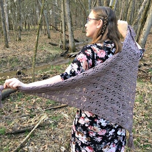 Puede incluir: Un chal triangular de color malva con un delicado diseño calado y borlas en los extremos. El chal se muestra al aire libre, resaltando su intrincado diseño y textura. Un accesorio hecho a mano.
