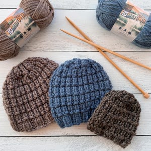 May include: Three knitted hats in brown and blue. The hats are made with a chunky yarn and have a textured stitch pattern. The yarn is in skeins in the background.
