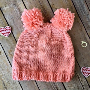 May include: A knitted coral-colored hat with two large pom-poms on top. The hat is made of a soft, textured yarn. Two heart-shaped patches and two small wooden rings are scattered around the hat on a wooden surface.