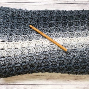 May include: A hand-crocheted blanket in grey and white hues, with a wooden crochet hook. The blanket displays a textured stitch pattern and a gradient effect, shifting from dark grey to light grey and white. The blanket's dimensions are not visible.