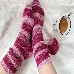 May include: A pair of pink and burgundy striped socks with a fuzzy texture. The socks are worn on a person's feet and are resting on a white fuzzy surface.