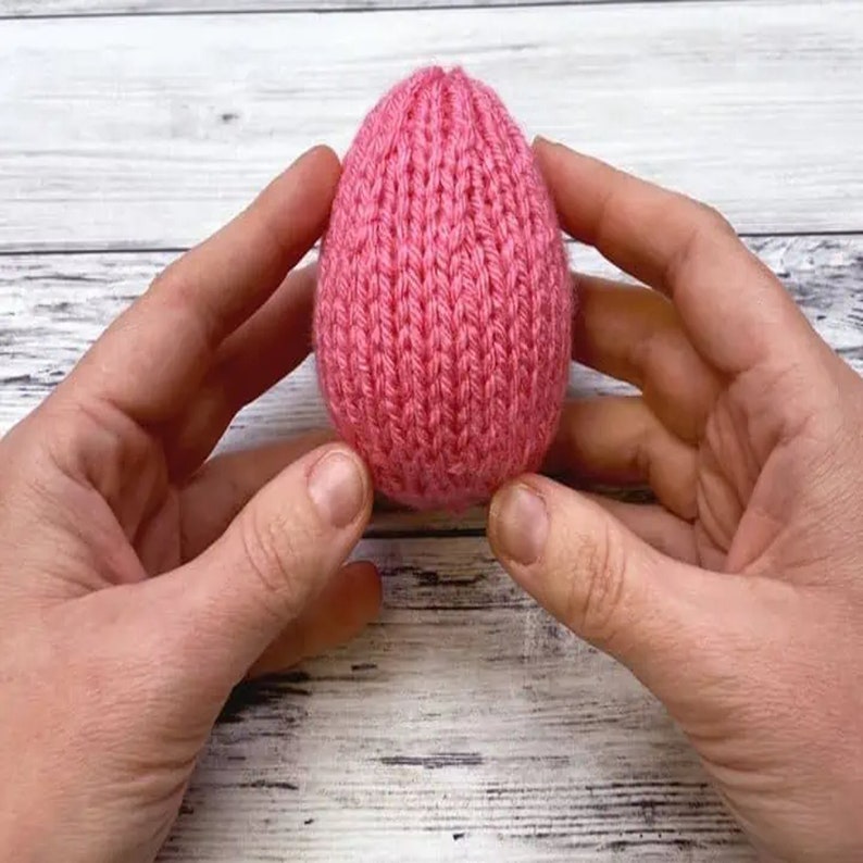 K&ouml;nnte beinhalten: Ein pinkfarbenes gestricktes Ei, das in zwei H&auml;nden gehalten wird.