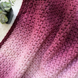 Könnte beinhalten: Eine handgehäkelte Decke in Rosa- und Burgundertönen. Die Decke hat ein strukturiertes Stichmuster mit einem Farbverlauf von Hellrosa zu tiefem Burgunder. Eine kleine Topfpflanze ist in der oberen linken Ecke zu sehen.