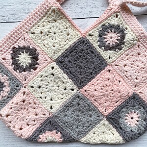 May include: A crocheted bag made of pink, white, and gray squares. The bag has a V-shaped opening and two straps.