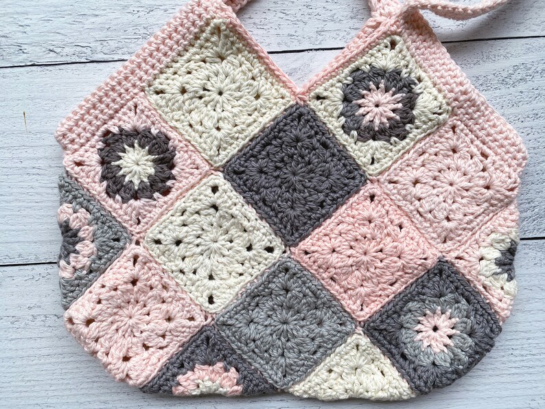 May include: A crocheted bag made of pink, white, and gray squares. The bag has a V-shaped opening and two straps.