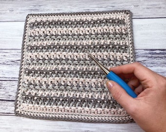 Puede incluir: Un cuadrado tejido a crochet con rayas alternas de hilo gris y rosa. Un gancho de crochet azul se sostiene en la mano derecha.