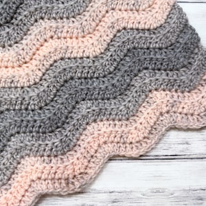 May include: A handmade crochet blanket in a wave pattern. The blanket features alternating bands of gray and light pink. The edges are scalloped. The blanket is made of soft yarn and is suitable for a baby or as a decorative throw.