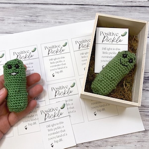 May include: A small crocheted green pickle with a smiling face and black eyes is in a wooden box with green moss. The box is next to a sheet of paper with four cards that say "Positive Pickle" and have a small pickle image on each card. The cards have text that says "Dill-ight in this little pickle, a reminder that you're kind of a big dill."