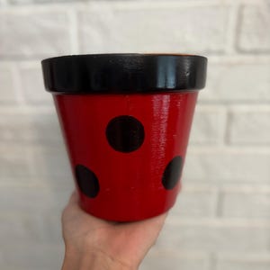 Puede incluir: Una maceta roja pintada con lunares negros y un borde negro. La maceta se sostiene frente a una pared de ladrillos blancos. La maceta está diseñada para parecerse a una mariquita.