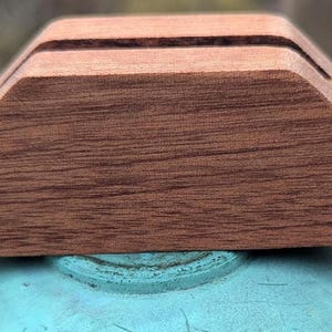 May include: A wooden phone stand with a triangular base and a slot for holding a phone. The stand is made of dark brown wood with a natural grain pattern.