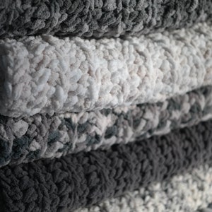 K&ouml;nnte beinhalten: Ein Stapel weicher, pl&uuml;schiger Decken in verschiedenen Grau- und Wei&szlig;t&ouml;nen. Die Decken haben ein dickes, strukturiertes Aussehen mit einem gem&uuml;tlichen, einladenden Aussehen. Die Farben reichen von Hellgrau bis Anthrazit und bilden eine neutrale Palette.