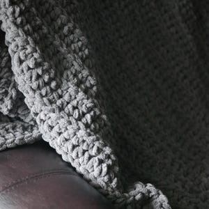 K&ouml;nnte beinhalten: Eine graue, grob gestrickte Decke, die &uuml;ber einer braunen Lederoberfl&auml;che drapiert ist. Die Decke hat eine weiche, pl&uuml;schige Textur mit sichtbaren Schlaufen und einer leicht unebenen Oberfl&auml;che. Die Farbe ist ein ged&auml;mpftes, neutrales Grau, das ein gem&uuml;tliches Gef&uuml;hl erzeugt.