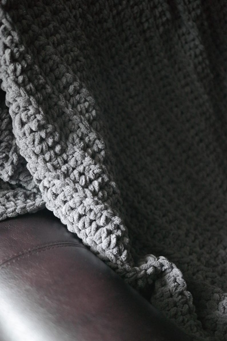 K&ouml;nnte beinhalten: Eine graue, grob gestrickte Decke, die &uuml;ber einer braunen Lederoberfl&auml;che drapiert ist. Die Decke hat eine weiche, pl&uuml;schige Textur mit sichtbaren Schlaufen und einer leicht unebenen Oberfl&auml;che. Die Farbe ist ein ged&auml;mpftes, neutrales Grau, das ein gem&uuml;tliches Gef&uuml;hl erzeugt.