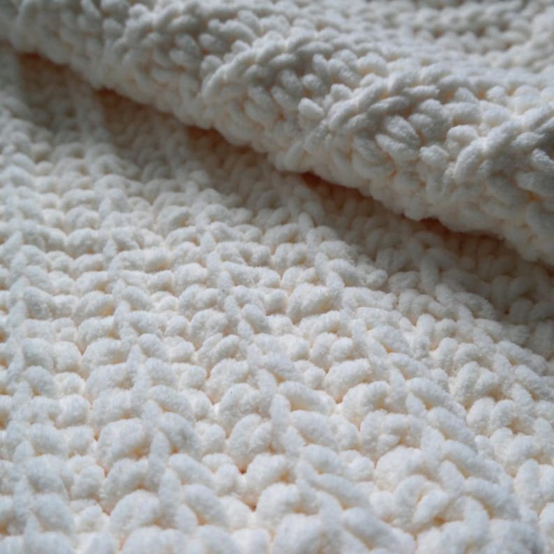 K&ouml;nnte beinhalten: Nahaufnahme einer cremefarbenen, grob gestrickten Decke. Der weiche, strukturierte Stoff ist gefaltet und zeigt die dicken, geschlungenen Maschen. Die Decke wirkt gem&uuml;tlich und warm, ideal f&uuml;r die Inneneinrichtung.