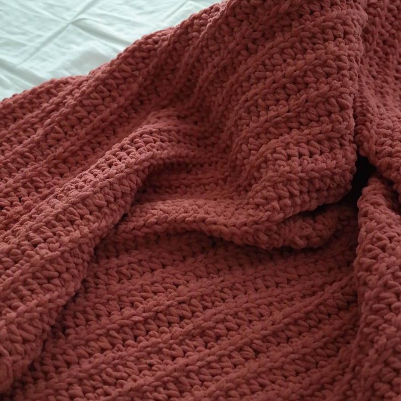 K&ouml;nnte beinhalten: Nahaufnahme einer handgefertigten, strukturierten Decke in einem ged&auml;mpften, staubigen Rosaton. Die Decke ist aus dickem Garn gefertigt, wodurch sie gem&uuml;tlich und warm wirkt. Die Stiche sind sichtbar und tragen zur handwerklichen &Auml;sthetik bei. Die Decke ist gefaltet und zeigt ihre Tiefe und Textur.