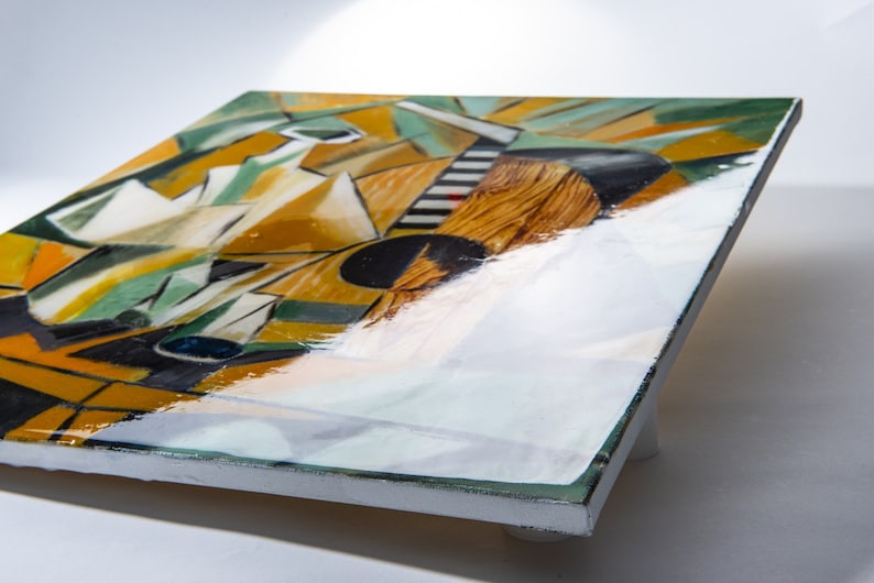 May include: A square canvas painting with a geometric abstract design in shades of yellow, green, black, and white. The painting has a glossy finish and is displayed on a white easel.