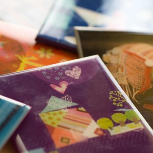 May include: A stack of colorful cards with different designs. The top card features a purple background with a white heart and a stylized house with a colorful roof. The card below it has a bright orange background with a floral design.
