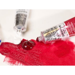 May include: A tube of Winsor & Newton Griffin Alkyd paint in a vibrant red color. The tube is lying on a white surface with a small amount of paint squeezed out.