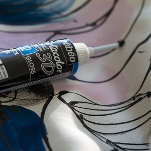 May include: A black and blue bottle of Pebeo Tacolor 3D gloss paint with a white cap and a silver applicator tip. The bottle is lying on a white fabric with black and blue abstract designs.