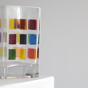 Pu&ograve; includere: Un bicchiere di vetro trasparente con un motivo geometrico colorato. Il motivo presenta quadrati di rosso, arancione, giallo, verde, blu e viola.