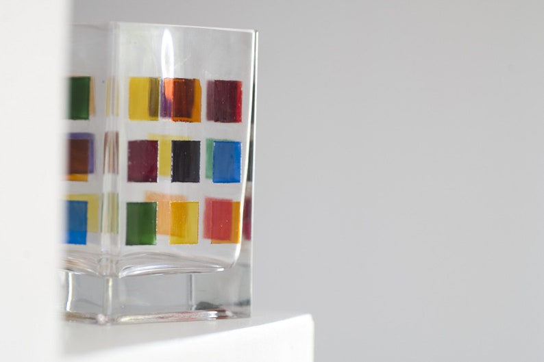 Pu&ograve; includere: Un bicchiere di vetro trasparente con un motivo geometrico colorato. Il motivo presenta quadrati di rosso, arancione, giallo, verde, blu e viola.