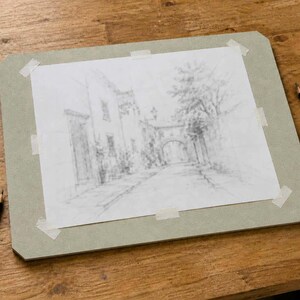 Peut inclure: Un croquis au crayon d'une sc&egrave;ne de rue est fix&eacute; sur une planche &agrave; dessin grise. Un taille-crayon, une gomme et plusieurs crayons sont dispos&eacute;s sur une surface en bois. Le croquis repr&eacute;sente des b&acirc;timents et une entr&eacute;e vo&ucirc;t&eacute;e.
