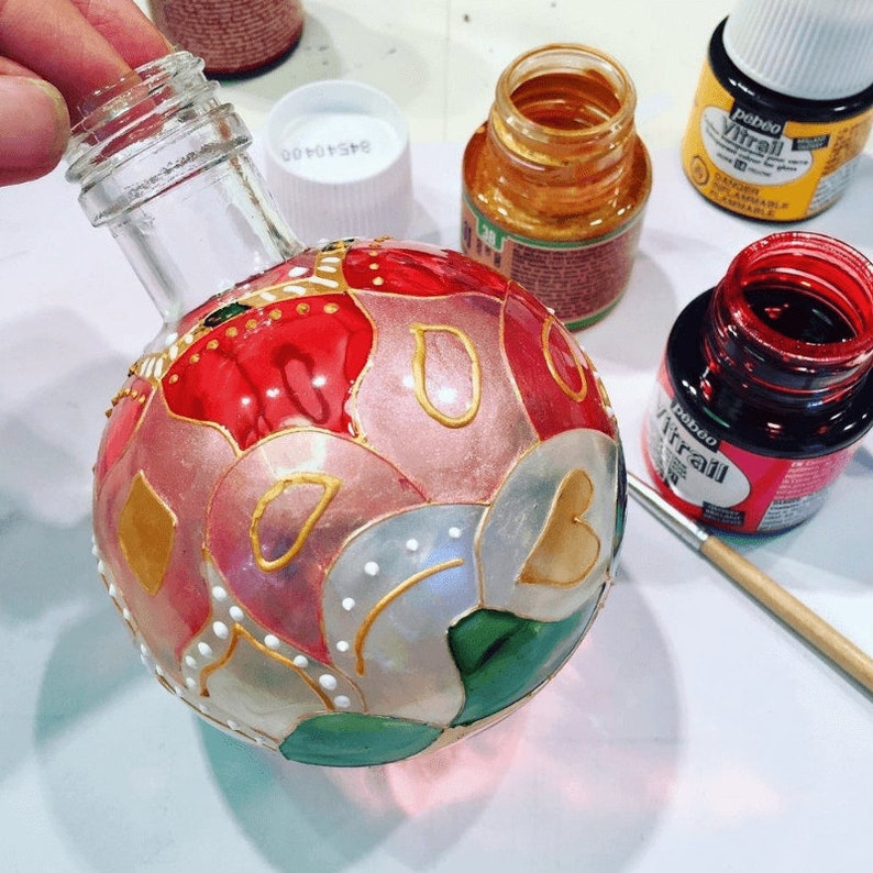 Pu&ograve; includere: Un addobbo natalizio in vetro con un motivo floreale dipinto in rosso, rosa, oro e verde. L'addobbo &egrave; in fase di decorazione con una piccola bottiglia di vernice dorata.