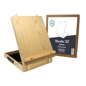 May include: A wooden pine box easel with a carrying handle and a built-in storage compartment. The easel is open and the lid is propped up at an angle. The easel is made by Studio 22 and is called the Hafren Pine Box Easel.