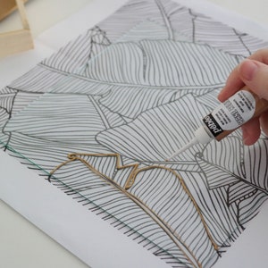K&ouml;nnte beinhalten: Eine Hand h&auml;lt eine Tube Goldfarbe und umrandet ein schwarz-wei&szlig;es Blattdesign auf einer Glasoberfl&auml;che.