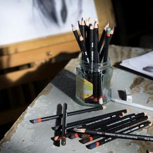 May include: A glass jar filled with black drawing pencils with red and gray colored pencils. The pencils are on a wooden table with a drawing pad and a wooden easel in the background.