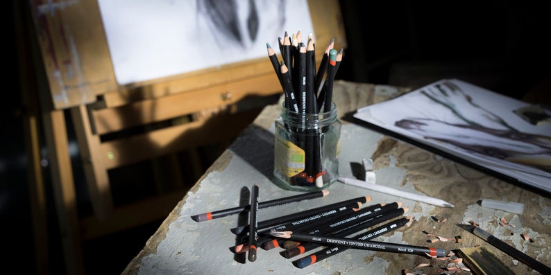 May include: A glass jar filled with black drawing pencils with red and gray colored pencils. The pencils are on a wooden table with a drawing pad and a wooden easel in the background.