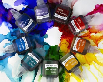May include: A set of ten 20 ml bottles of alcohol ink in various colors, including ultramarine, lake blue, turquoise, cobalt green, amber yellow, orange, cadmium red, crimson, and lavender. The bottles are arranged in a circle on a white surface with colorful alcohol ink splatters.